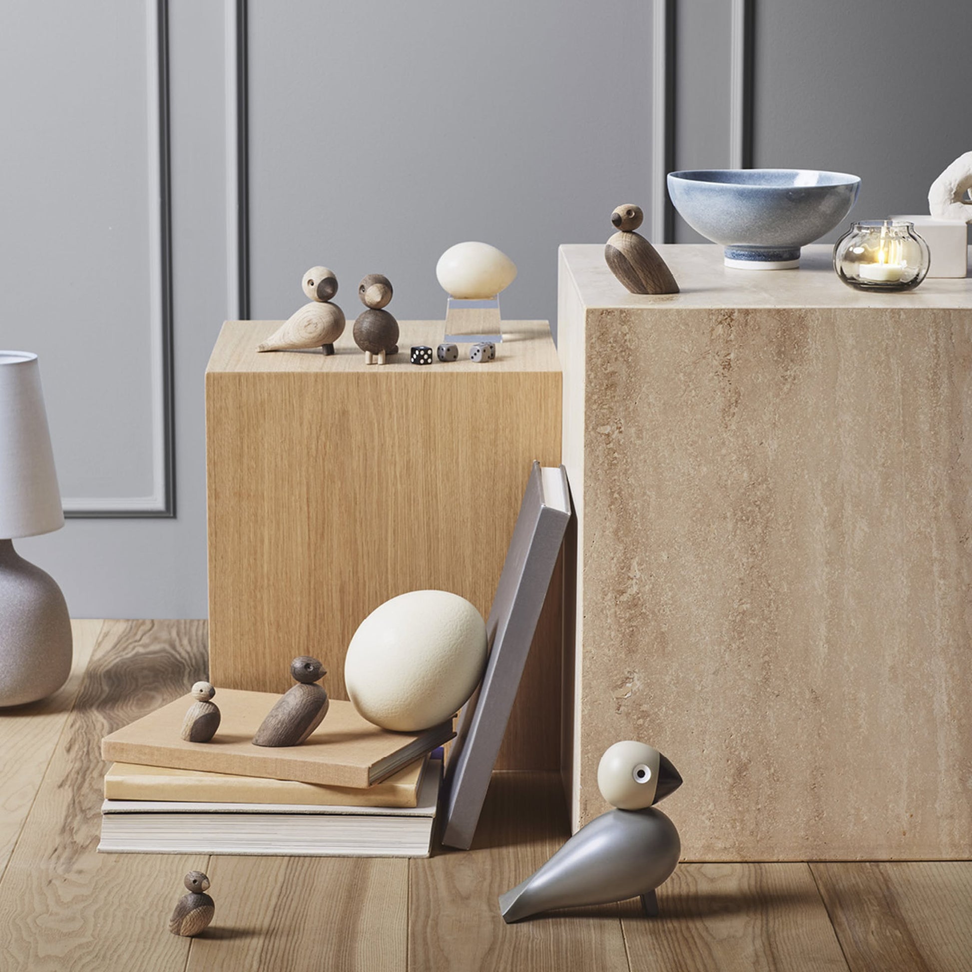 A minimalist scene featuring the Kay Bojesen Lovebirds Pair by Kay Bojesen displayed on wooden cubes, books, and the floor; styled with a lamp, bowl, candle, and neutral decor against a gray paneled wall.