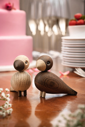 The Kay Bojesen Lovebirds Pair by Kay Bojesen stands on a table before a pink cake, stacked white plates, strawberries, and wine glasses, adding festivity and elegance to the setting with their Danish craftsmanship.