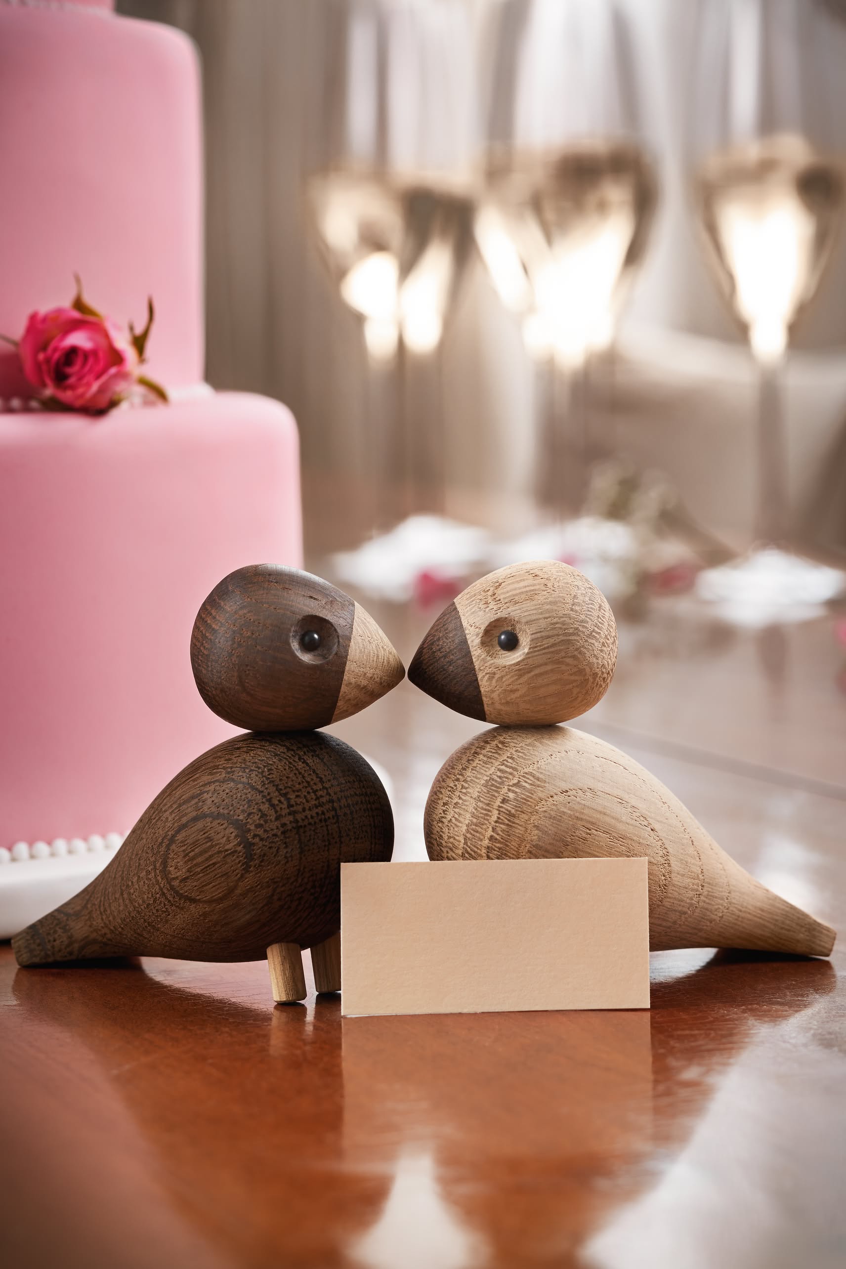 The Kay Bojesen Lovebirds Pair by Kay Bojesen face each other on a reflective table with a blank card between them. A pink cake and wine glasses in the background evoke Danish craftsmanship and a romantic atmosphere.