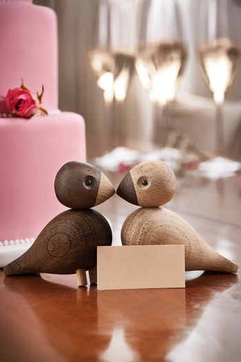 The Kay Bojesen Lovebirds Pair by Kay Bojesen face each other on a reflective table with a blank card between them. A pink cake and wine glasses in the background evoke Danish craftsmanship and a romantic atmosphere.