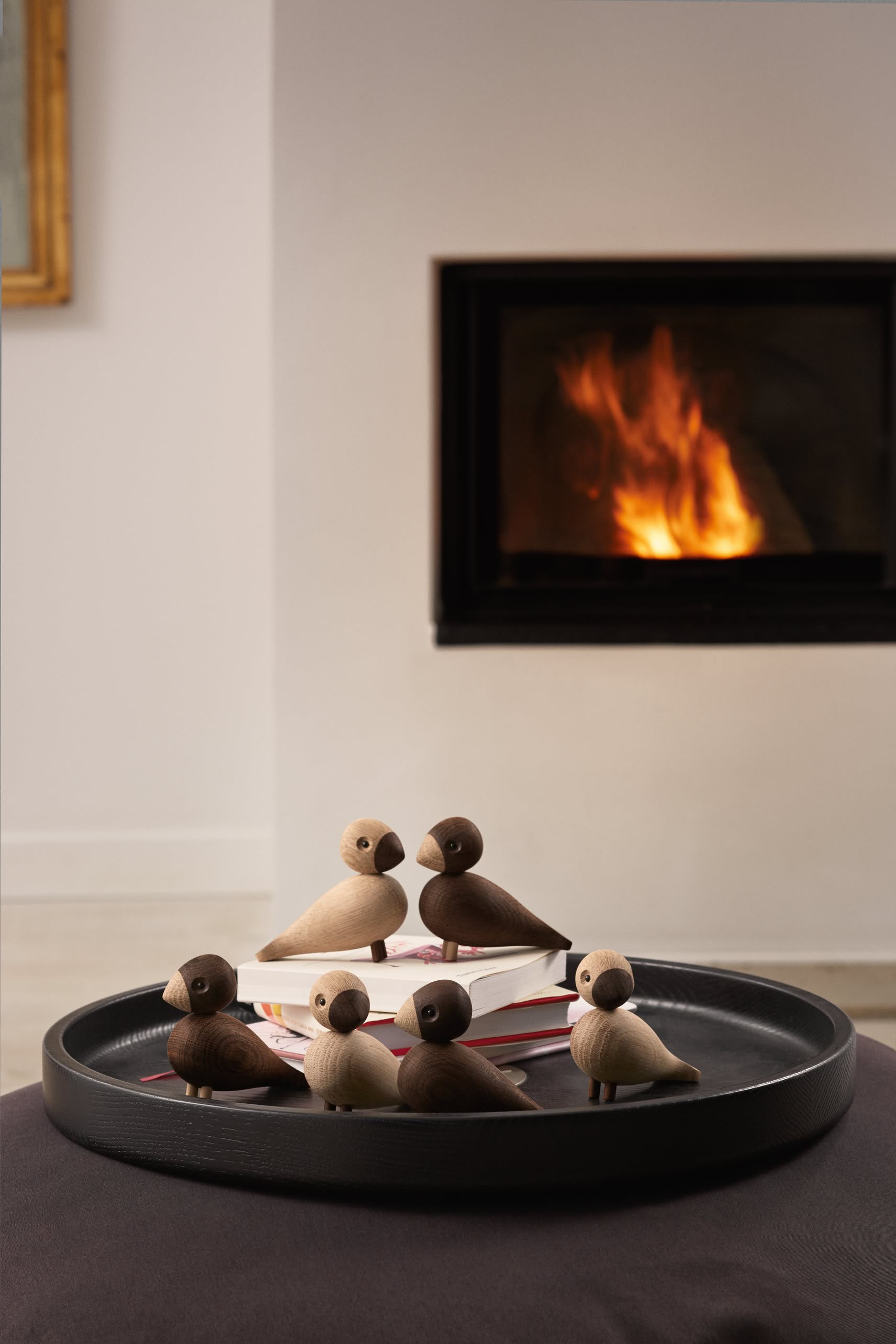 A black round tray with the Kay Bojesen Lovebirds Pair by Kay Bojesen and books rests on a dark surface before a lit fireplace, creating a cozy indoor scene.
