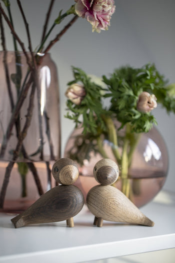 The Kay Bojesen Lovebirds Pair by Kay Bojesen sits on a white surface before pink glass vases with flowers and foliage, creating a soft decorative scene that celebrates Danish craftsmanship.