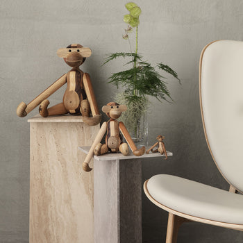 The Kay Bojesen Monkey Mini from Kay Bojesen is displayed on a stone pedestal beside a modern white chair, with a green leafy plant in a clear vase and gray wall in the background—a delightful collectible keepsake.