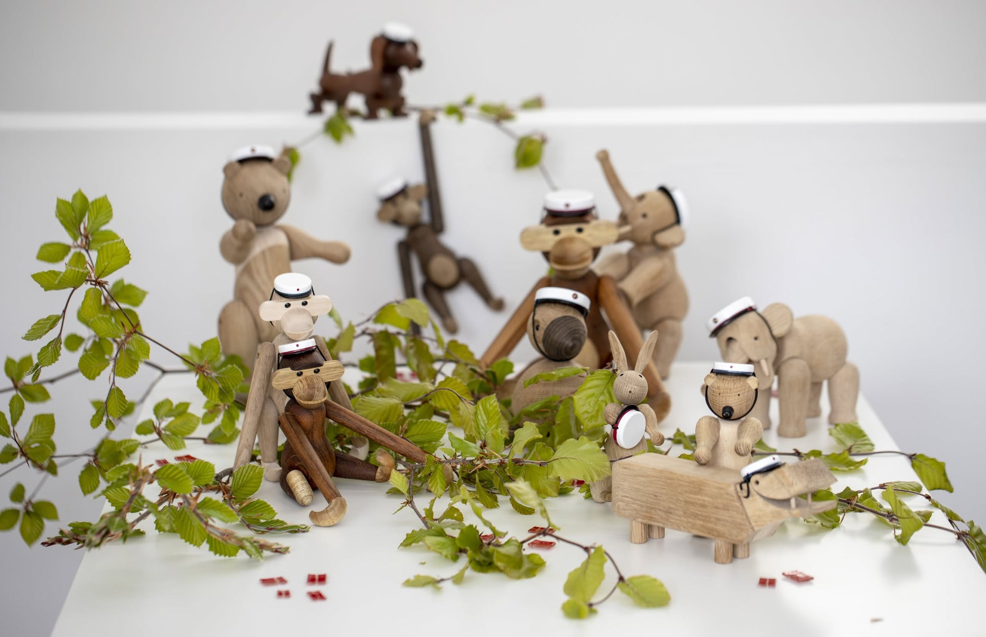 A group of Kay Bojesen wooden figurines, including the Kay Bojesen Monkey Small, each with a white graduation cap, is arranged on a white surface decorated with green leafy branches.