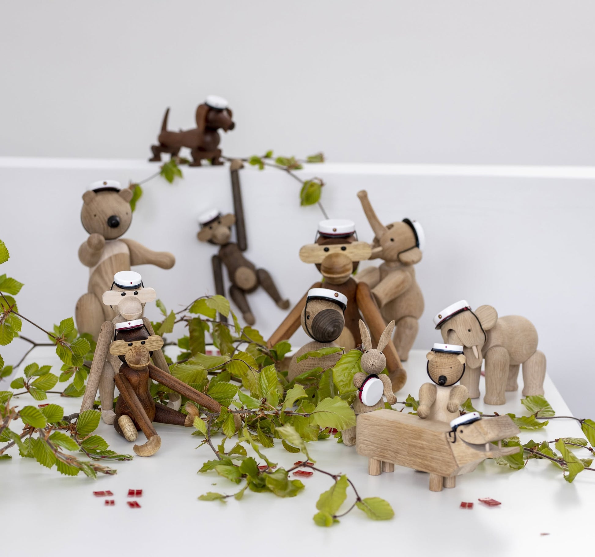 The Kay Bojesen Monkey Small by Kay Bojesen, wearing a white graduation cap, is displayed on a white surface with green leaves. This classic wooden figurine stands out against the light background.