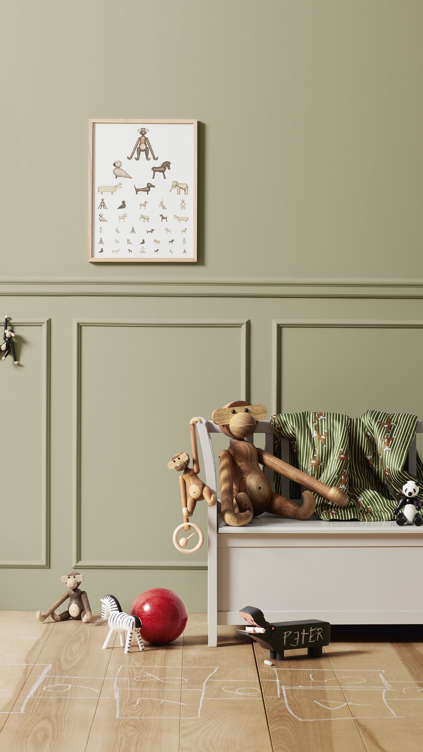 A children’s room with green walls, a framed animal poster, a white bed with striped pillows, stuffed animals—including the Kay Bojesen Monkey Small by Kay Bojesen—a red ball, zebra toy, and chalk drawings on the wooden floor.