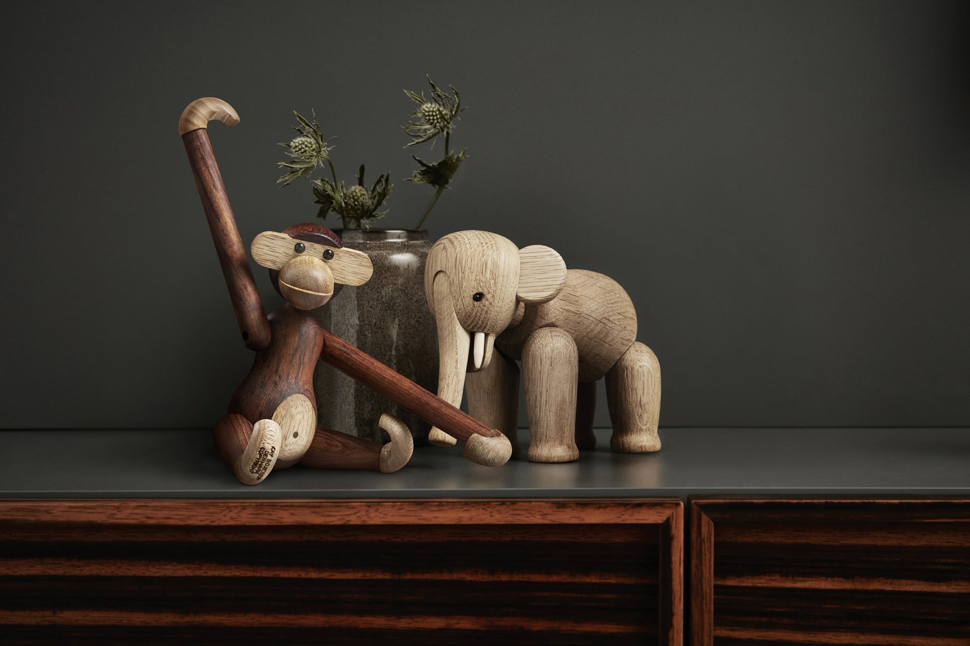 The Kay Bojesen Monkey Small by Kay Bojesen sits with one arm raised beside a wooden elephant on a dark surface, with a glass vase of dried flowers in the background against a dark wall.