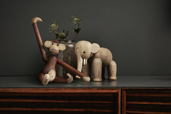 The Kay Bojesen Monkey Small by Kay Bojesen sits with one arm raised beside a wooden elephant on a dark surface, with a glass vase of dried flowers in the background against a dark wall.