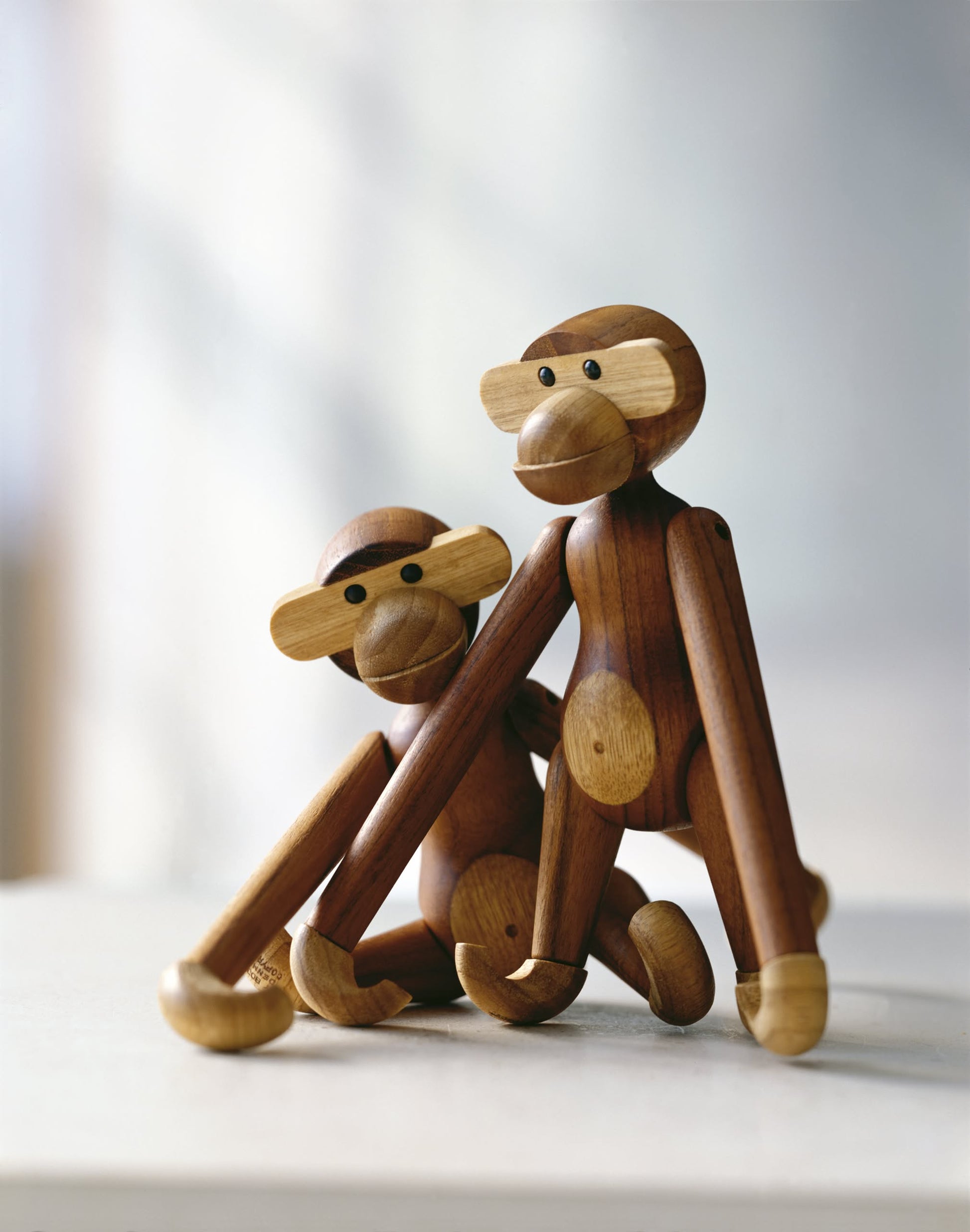 Two Kay Bojesen Monkey Small figurines by Kay Bojesen, with long arms, sit side by side on a light surface. One leans forward while the other sits upright—an iconic and timeless design piece for any space.