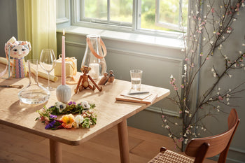 A wooden table by a sunlit window holds a flower wreath, a glass of water, a candle, Kay Bojesen’s iconic Monkey Small by Kay Bojesen, glassware, a notepad, and a whimsical cat-shaped pitcher. A blooming branch stands nearby.