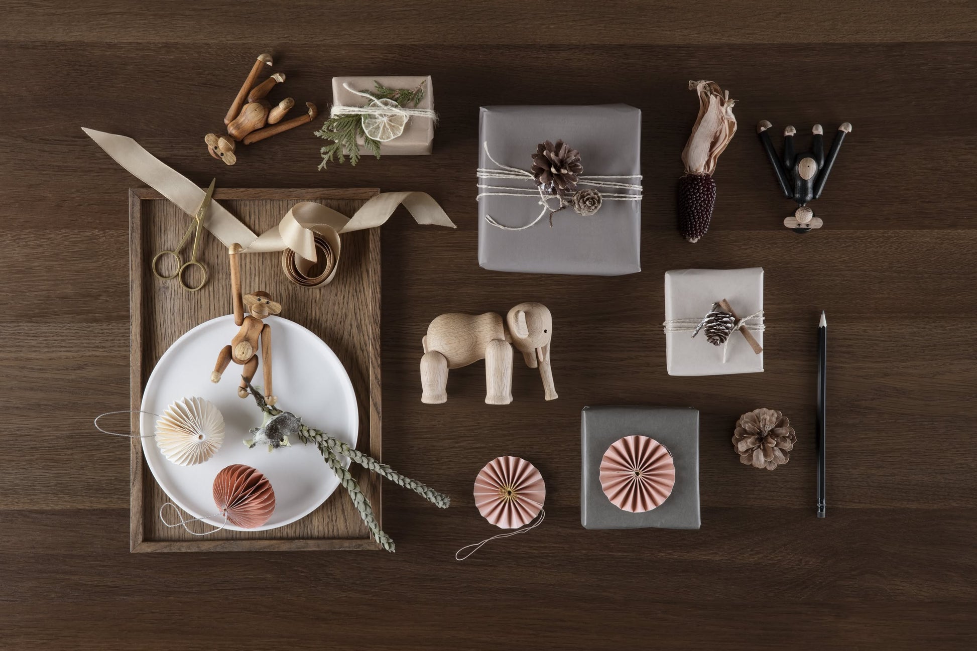 A flat lay on a wooden surface features wrapped gifts with natural decorations, the timeless Kay Bojesen Monkey Small by Kay Bojesen, wooden dolls, rings on a plate, dried plants, paper ornaments, a pencil, and a pinecone.