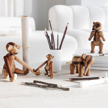 The Kay Bojesen Monkey Small by Kay Bojesen is arranged with other wooden animal figurines on a light wood table featuring twig pencils, rolled paper, and books. A blurred white sofa appears in the background.