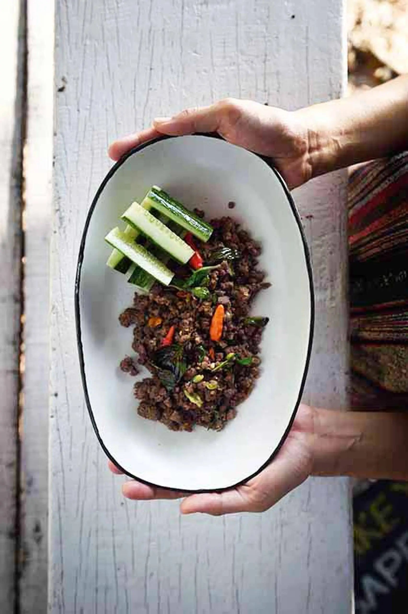 A person holds a Kra Sanctuary Andaman Family Sharing Dish by Kra Sanctuary, its classic white glaze filled with stir-fried minced meat, basil, chilies, herbs, and cucumber sticks on a rustic white wooden surface inspired by island life.