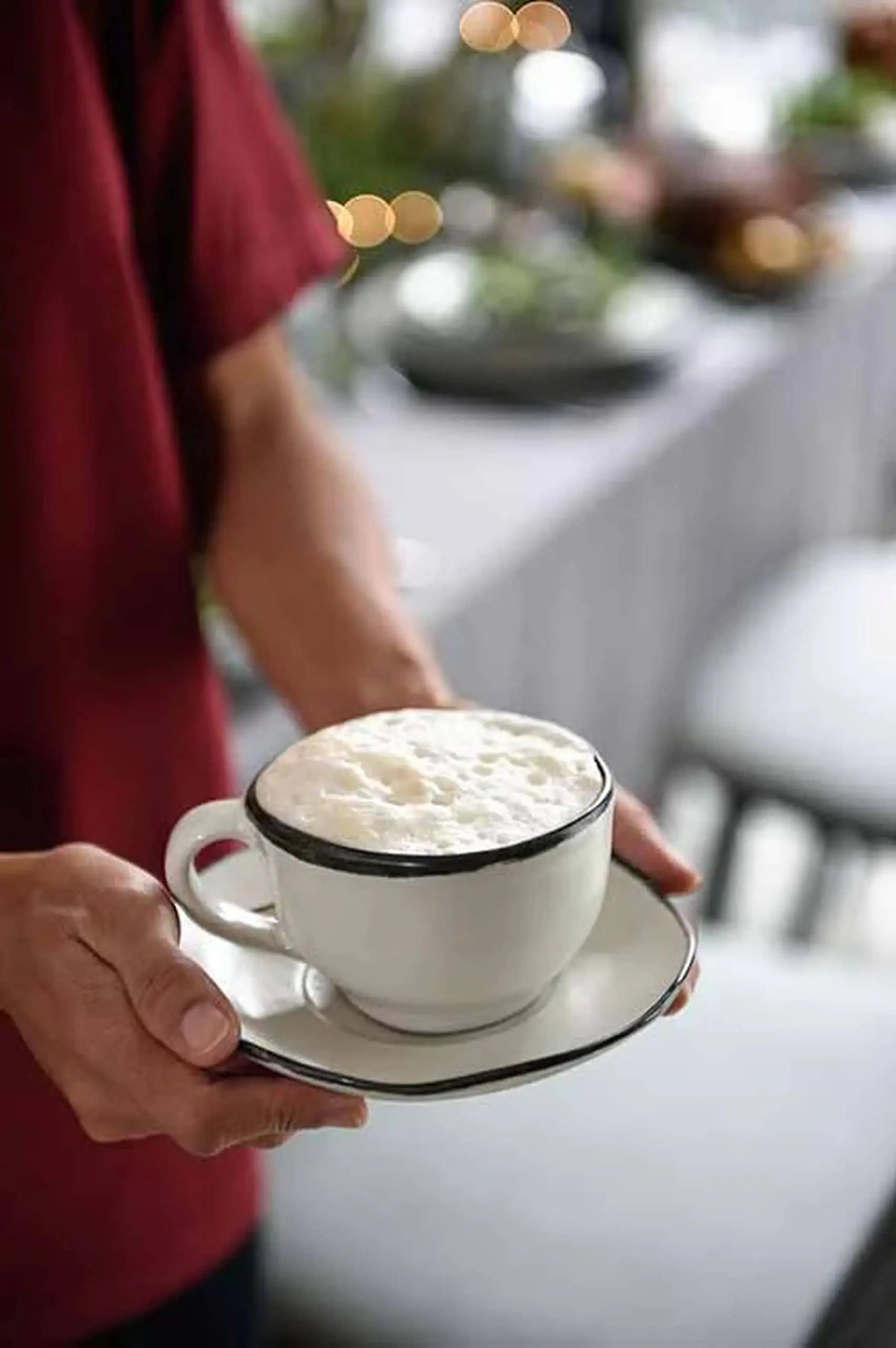 A person in a red shirt holds a Kra Sanctuary Andaman Free-Form Cup and Coaster, filled with frothy coffee, against a blurred backdrop of diningware and chairs—perfectly capturing the essence of island living.