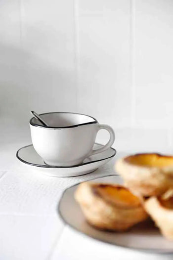 A Kra Sanctuary Andaman Free-Form Cup and Coaster, with a spoon inside, sits on a white surface. In front, a plate of blurred egg tarts appears, set against a bright, minimalistic background evoking island living.