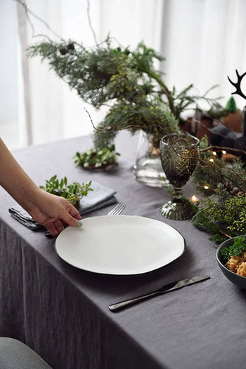 A hand sets the Kra Sanctuary Andaman Free-Form Dinner Plate on a gray tablecloth, surrounded by silverware and a green plant centerpiece for an elegant, festive island living table setting.