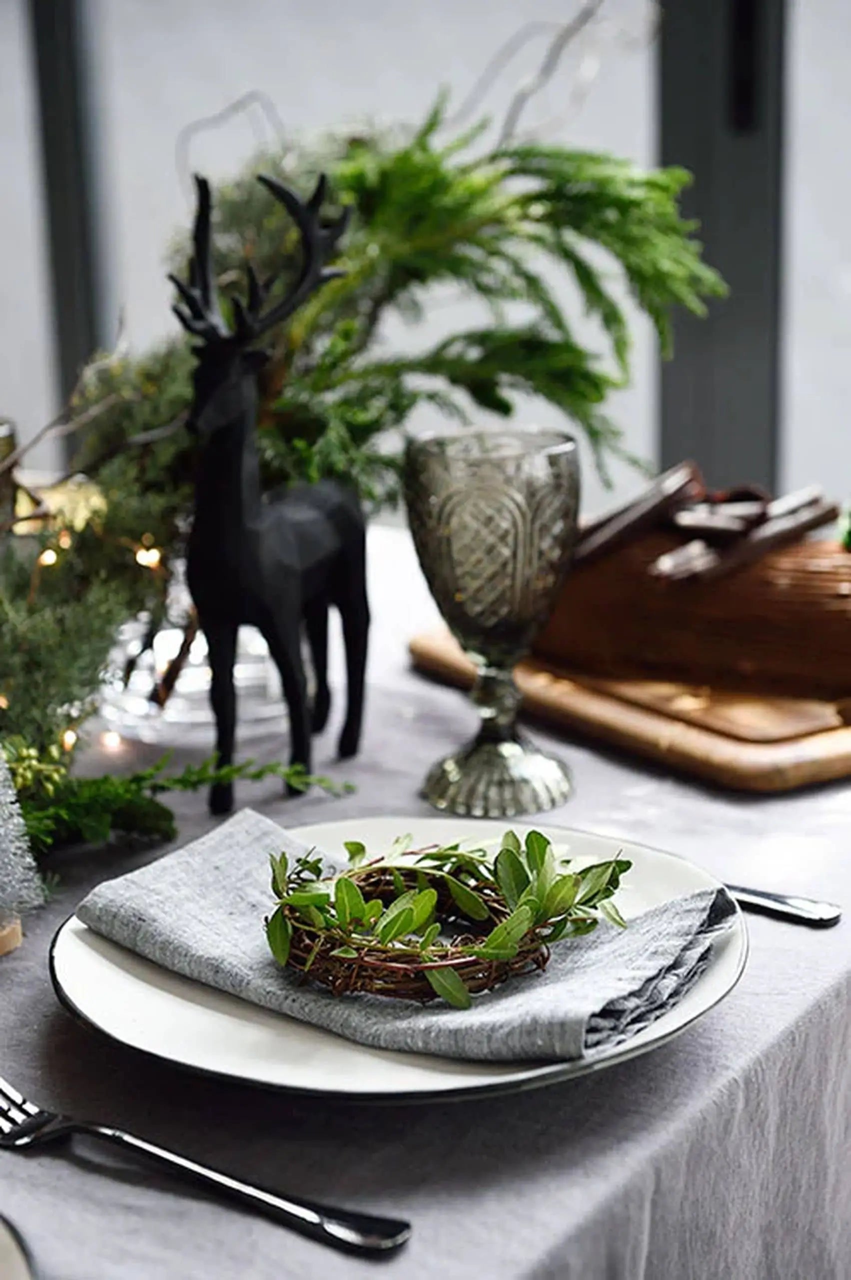 A festive table is set with the Kra Sanctuary Andaman Free-Form Dinner Plate, classic white glaze, paired with a gray napkin and leafy wreath. A black deer figurine, lush greenery, and a patterned goblet evoke elegant island living.