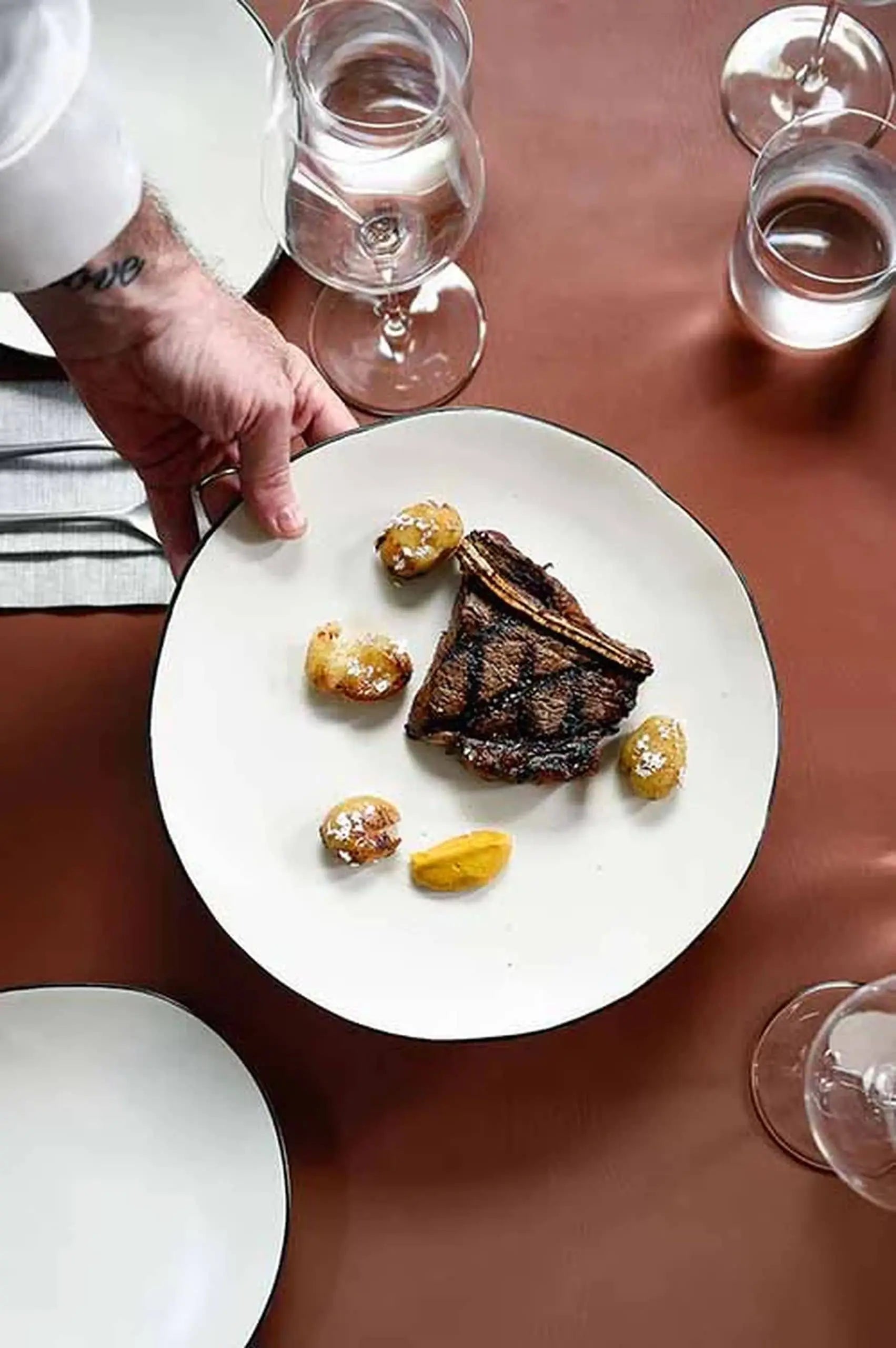 A person serves grilled steak, roasted baby potatoes, and a lemon wedge on the Kra Sanctuary Andaman Free-Form Dinner Plate. The setting recalls island living with wine glasses, water glasses, and a brown tablecloth.