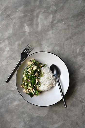 White rice and mixed green vegetable stir-fry are served in the Kra Sanctuary Andaman Free-Form Pasta Bowl by Kra Sanctuary, set on a gray surface with a fork and spoon on either side.