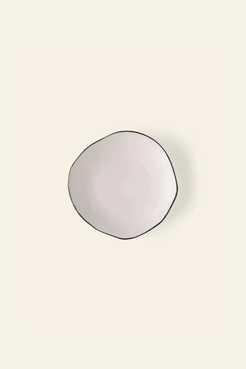 The Kra Sanctuary Andaman Free-Form Side Plate by Kra Sanctuary is a round, off-white ceramic plate with an irregular shape and thin dark rim, reflecting relaxed island elegance against a light beige background.