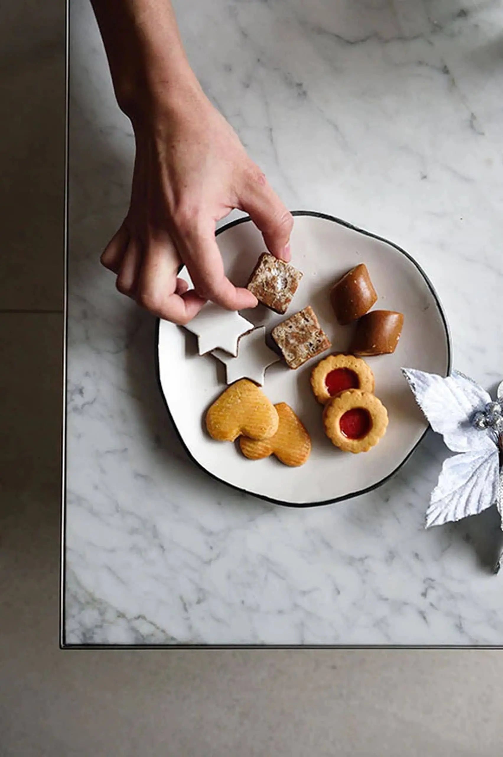 A hand reaches for a star-shaped cookie from a Kra Sanctuary Andaman Free-Form Side Plate by Kra Sanctuary on a marble table beside a silver poinsettia, evoking the warmth and charm of an Andaman-inspired dining experience.