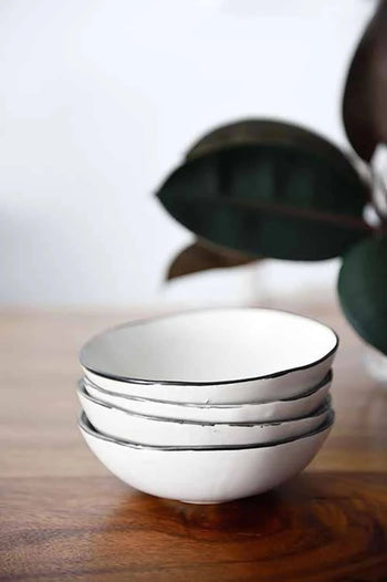 Three Kra Sanctuary Andaman Free-Form Soup Bowls in classic white ceramic are stacked on wood, with green leaves softly blurred in the background. These bowls add a minimal, serene touch to your dining and perfectly complement any Andaman Diningware collection.