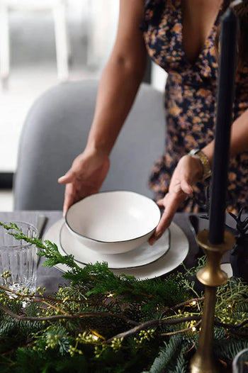 A person in a floral dress sets a Kra Sanctuary Andaman Free-Form Soup Bowl on a matching plate at a decorated table with greenery, glassware, and a black candle in brass, evoking the relaxed elegance of island living.