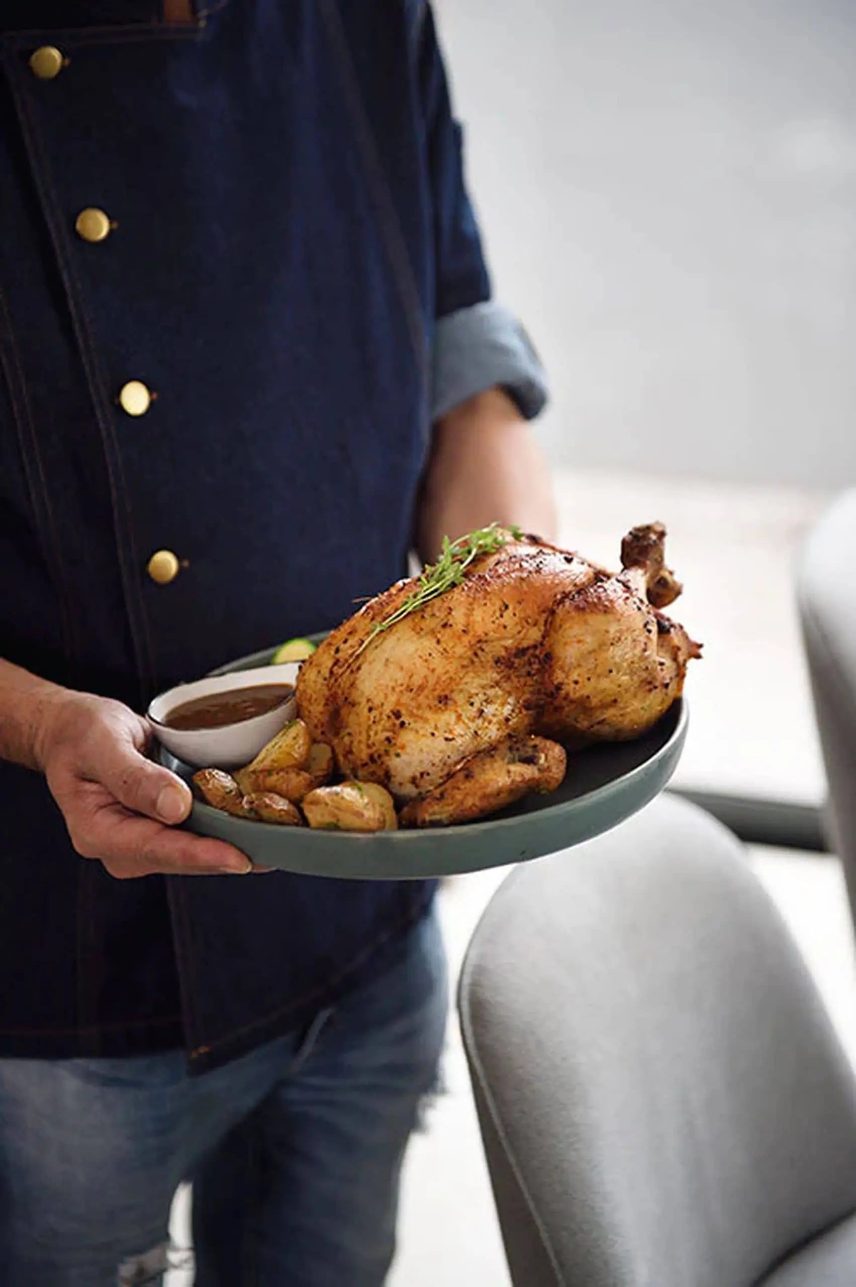 A person in a dark buttoned shirt holds a plate with a whole roasted chicken, small potatoes, and herbs, served on the Kra Sanctuary Karst Flat Dinner Plate by Kra Sanctuary for a unique look. A small bowl of sauce on the side completes the rustic charm.