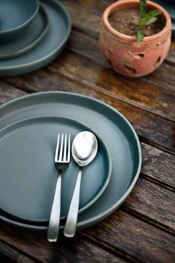 The Kra Sanctuary Karst Flat Side Plate is styled with a fork and spoon on a rustic wooden table beside a clay pot with a small plant, adding charm to your dining setting.