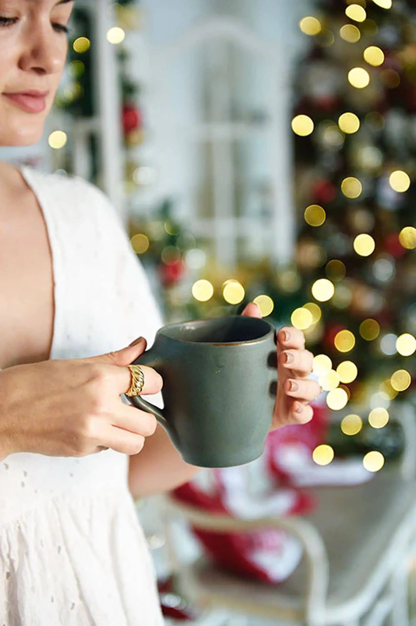 Someone in a white top holds the Kra Sanctuary Karst Free-Form Coffee Mug by Kra Sanctuary, with a decorated Christmas tree and festive lights softly blurred in the background for a unique look.