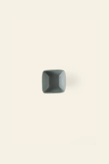 The Kra Sanctuary Karst Square Snack Bowl, a small empty gray bowl, is centered on a light beige background, casting a faint shadow to the right and highlighting its distinctive design.