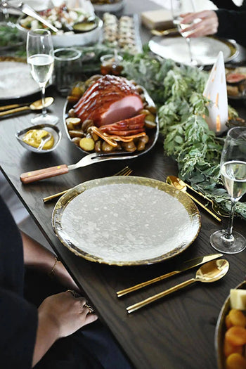 A festive table set with gold cutlery, Kra Sanctuary Satay Free-Form Dinner Plates, wine glasses, greenery decor, and a platter of roasted meat with potatoes in the background.