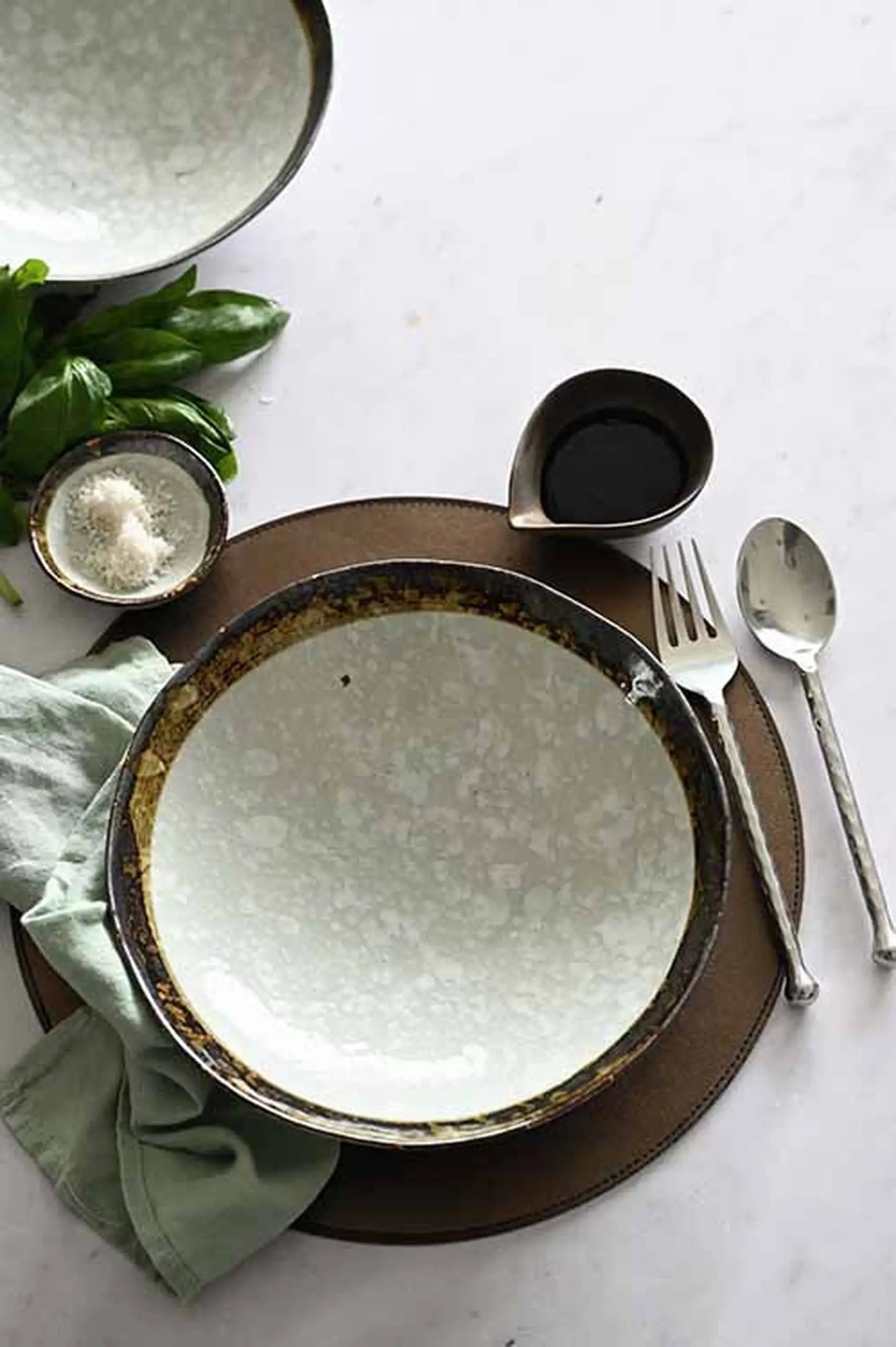The Kra Sanctuary Satay Free-Form Pasta Bowl by Kra Sanctuary rests on a brown placemat with cutlery, a small dark liquid pitcher, grated cheese, herbs, and a green napkin—ideal for creating an inviting dining experience.