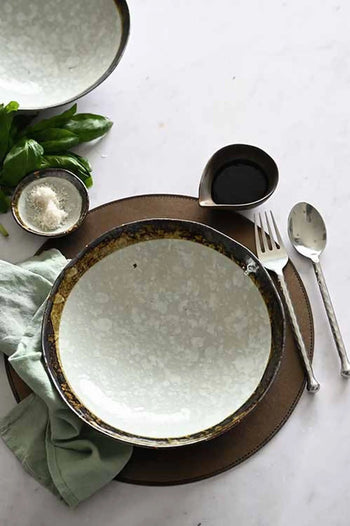 The Kra Sanctuary Satay Free-Form Pasta Bowl by Kra Sanctuary rests on a brown placemat with cutlery, a small dark liquid pitcher, grated cheese, herbs, and a green napkin—ideal for creating an inviting dining experience.