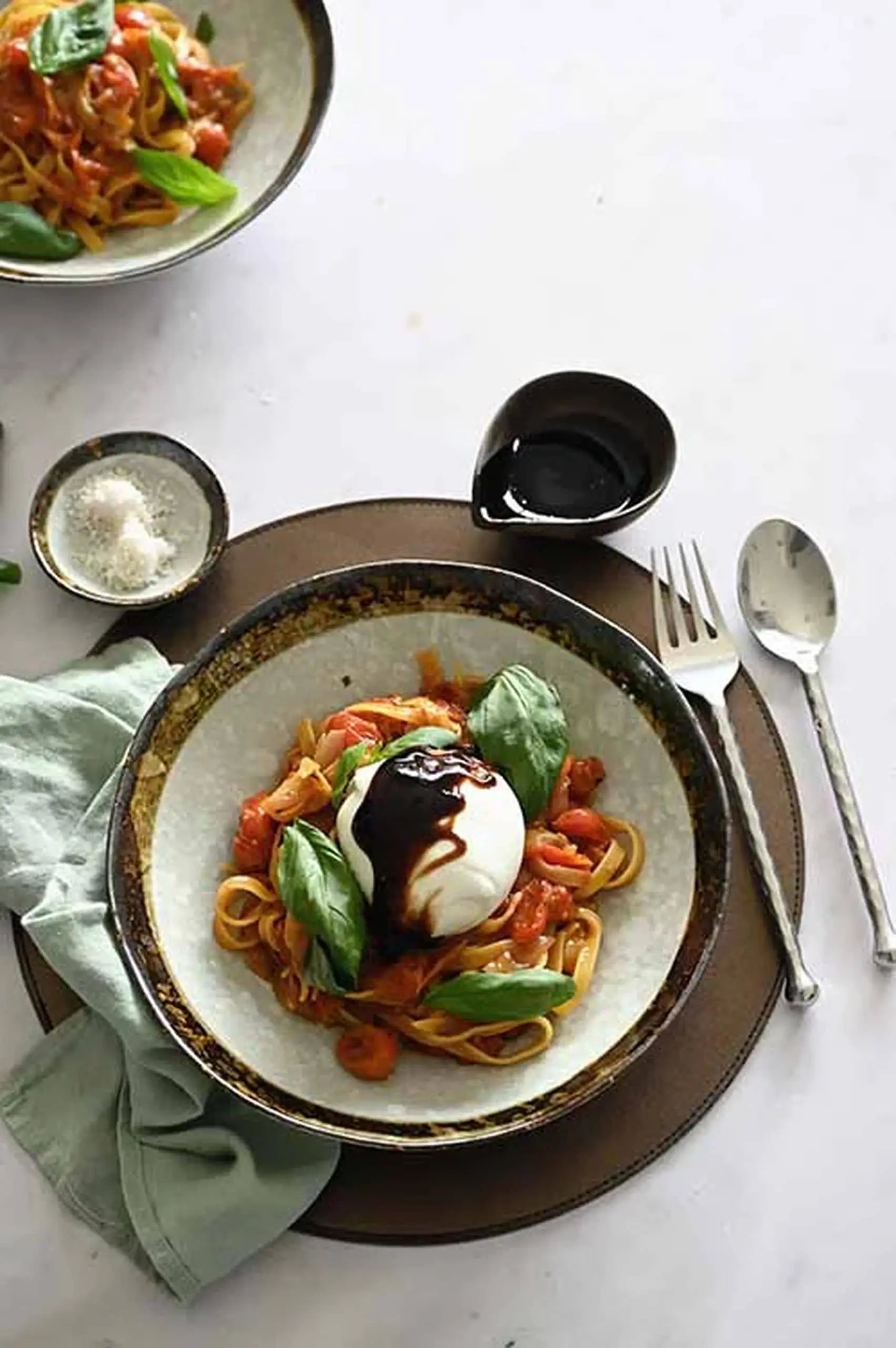 A serving of pasta with tomato sauce, basil, and burrata in the Kra Sanctuary Satay Free-Form Pasta Bowl by Kra Sanctuary, set with a green napkin, fork, spoon, plus small dishes of salt and balsamic for extra flavor.