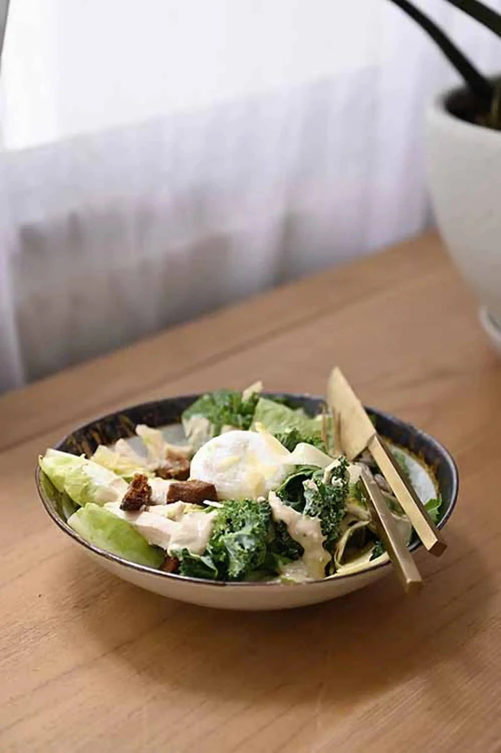 A Caesar salad with lettuce, croutons, parmesan, and creamy dressing is served in a Kra Sanctuary Satay Free-Form Pasta Bowl by Kra Sanctuary on a wooden table next to a gold fork and knife. A potted plant is softly blurred in the background near the window.