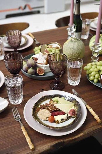 A wooden dining table set with Kra Sanctuary Satay Free-Form Side Plates, assorted cheeses, crackers, figs, grapes, wooden-handled cutlery, a melon, glassware, and tall pink candles for an elegant dining experience.