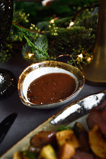 The Kra Sanctuary Satay Shell Dipping Dish by Kra Sanctuary, a rustic ceramic bowl with an iridescent border, holds brown gravy on a dark table amid festive greenery and string lights, with a nearby plate of food partially visible.