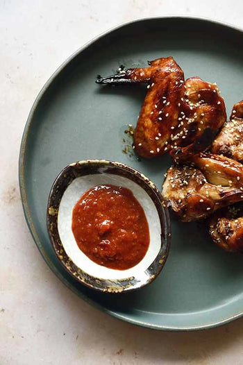 Glazed chicken wings with sesame seeds rest on earthy dinnerware, accompanied by a Kra Sanctuary Satay Shell Dipping Dish from Kra Sanctuary filled with reddish sauce.