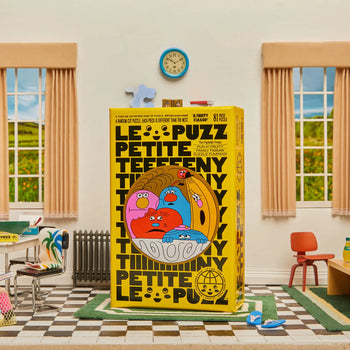 Le Puzzs A Fruity Fiasco 81-piece puzzle box, featuring bold black text and a colorful fruit bowl, is displayed at the center of a retro room with two windows, a clock, beige curtains, and classroom furniture.