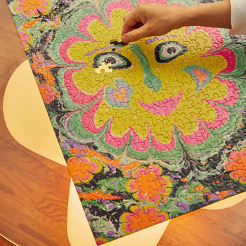 A hand places the last piece into Le Puzz Freaky Deaky, a 500-piece puzzle by Le Puzz, showing a vibrant, stylized flower with two eyes at its center on a wooden table with yellow decorations.