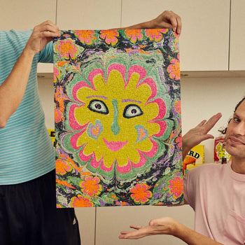 Two people display the completed Le Puzz Freaky Deaky 500 Piece Puzzle by Le Puzz, featuring an abstract flower with a smiling face and a vibrant swirl of pink, yellow, green, and orange in the background.