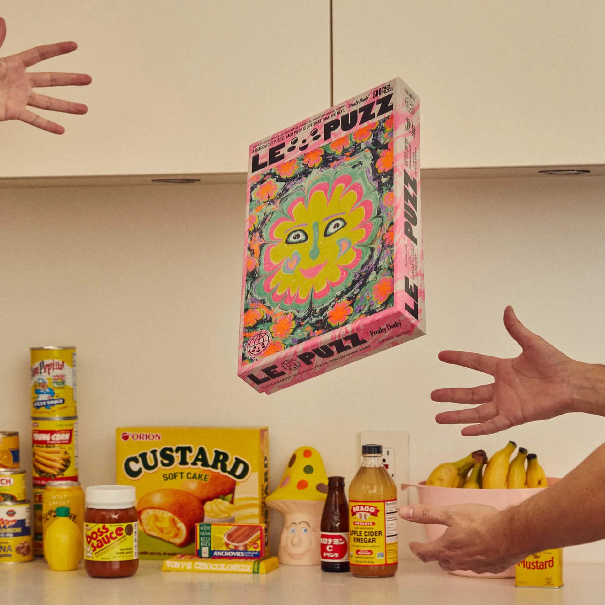 The Le Puzz Freaky Deaky 500 Piece Puzzle by Le Puzz floats in midair between two hands above a kitchen counter with custard, mustard, fruit, and groceries including a banana bunch for a unique experience.