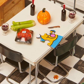 A whimsical tabletop scene: a pumpkin, wine and glasses, celery, packaged meat, a small knife, and the Le Puzz Head in the Clouds 81 Piece Puzzle by Le Puzz. Black chairs on a checkered floor—perfect for sparking imagination.