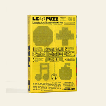 A yellow Le Puzz Kindness is Contagious 500 Piece Puzzle box with black text and graphics, showing instructions for creating your own pearler beads and pixel art designs like a heart, circle, cross, and music note on the front.