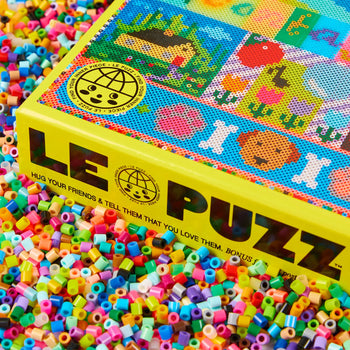 A bright yellow Le Puzz Kindness is Contagious, 500 Piece Puzzle box sits on colorful beads, featuring playful dot art and a smiling globe with a message spreading hugs and love to friends.