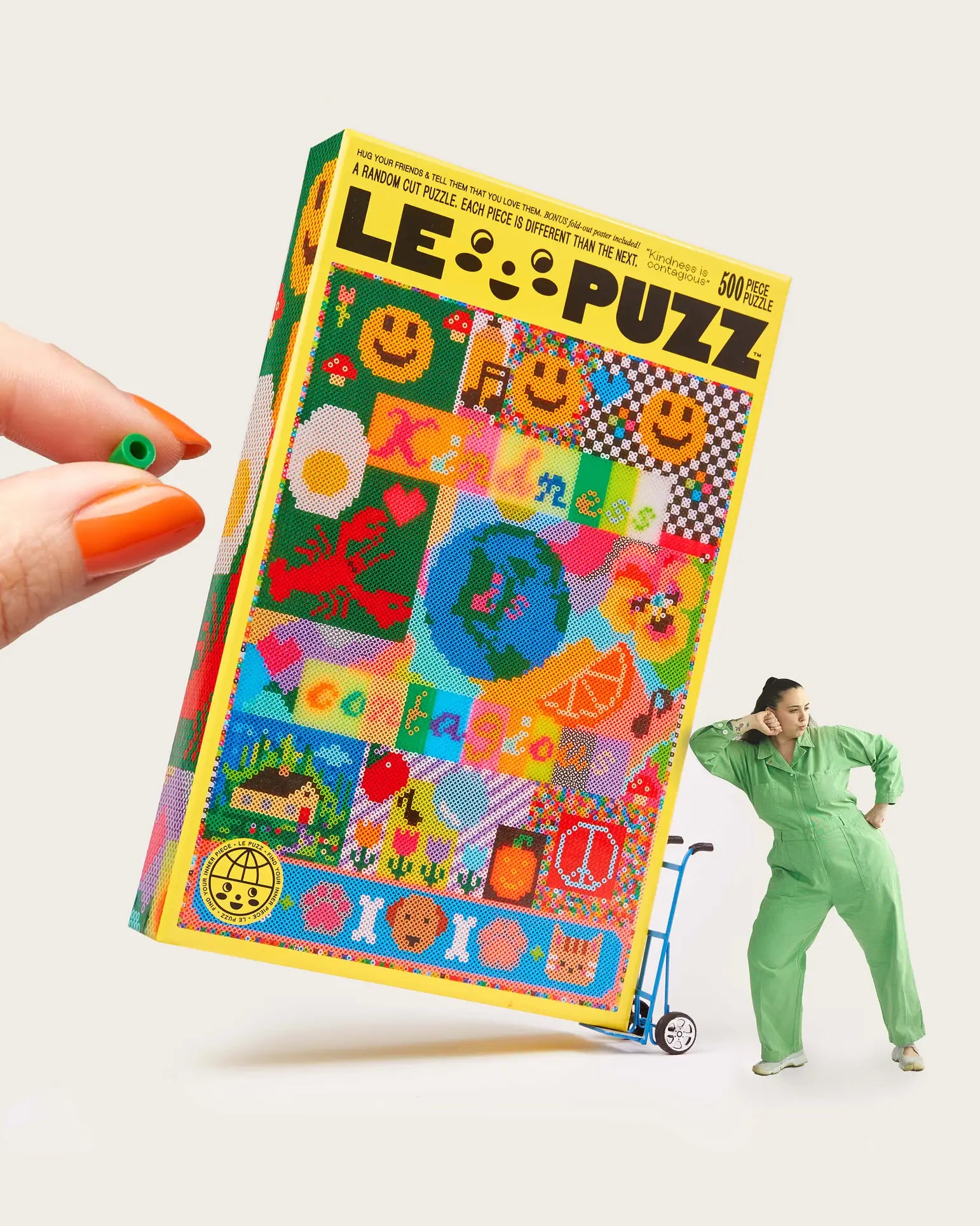 A hand with orange-painted nails holds the Le Puzz Kindness is Contagious 500 Piece Puzzle box as a person in a green jumpsuit leans on a blue hand truck beside the oversized Le Puzz box.
