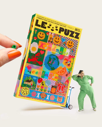A hand with orange-painted nails holds the Le Puzz Kindness is Contagious 500 Piece Puzzle box as a person in a green jumpsuit leans on a blue hand truck beside the oversized Le Puzz box.