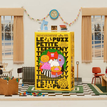 A bright yellow Le Puzz The Flumpsteins 81 Piece Puzzle by Le Puzz sits at the center of a lively classroom, surrounded by scattered toys, chairs, and art supplies. Cream walls, two large windows, and a clock complete the scene.