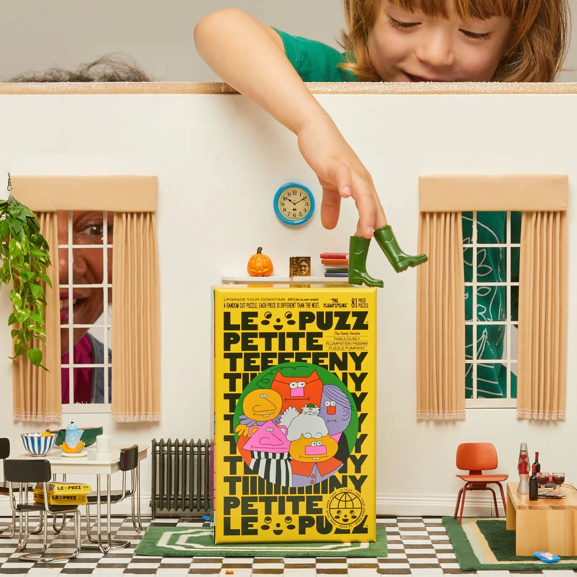 A child balances a green boot on the Le Puzz The Flumpsteins, 81 Piece Puzzle box in a vibrant room with playful furniture, plants, and Flumpsteins decor as an adult peeks through the window—ready for new adventures.